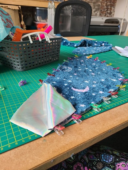 A sewing table with cutting mats, showing a sandwich of 3 wing-shaped pieces of fabric. They are clipped together, and the top two layers are folded at the end to show iridescent spandex inside. The top layer is a space-themed soft minky.