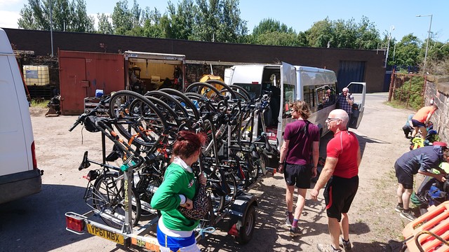 Lots of bikes packed onto a trailer which is hooked up to a minibus, with a few cyclists milling around it doing final checks 