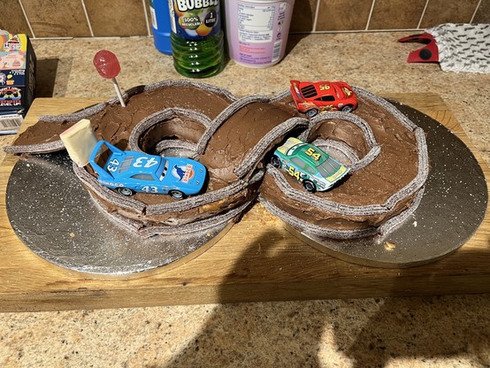 A figure of 8 race track made out of two interlocking ring cakes. It’s covered in chocolate icing and sweets to represent the signs. A bridge completes the 8 and a ramp leads on to the track. Three cars, all characters from the movie Cars, race around the track. 