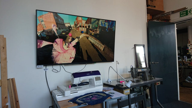 View of a room in a makerspace.  A CNC embroidery machine and an industrial sewing machine are on tables next to the wall.

Above them is a huge TV mounted on the wall, it's almost the same size as the door to the room, which is visible off to the right.

The TV is showing a video of a person dressed in a pink costume with pink crocheted hat with rabbit ears, gesturing towards an old warehouse building in a gritty urban street