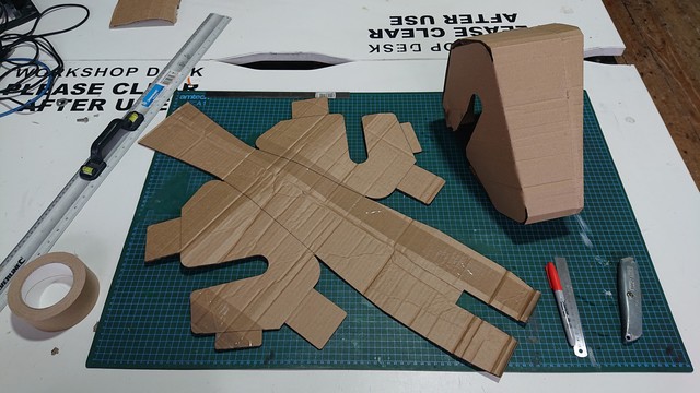 A workbench with a cutting mat, some tape, a sharpie, a Stanley knife and a metal ruler.  On the cutting mat is a flat sheet of brown corrugated cardbaord cut out in the shape of the unwrapped mesh from the CAD design.  Next to that is a version which has been folded up to make the triangular wedge shape.