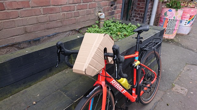 A gravel bike with drop handlebars leans against a planter outside a brick building.  On the handlebars, looking a bit like a head for the bike, is the assembled brown cardboard wedge shape.