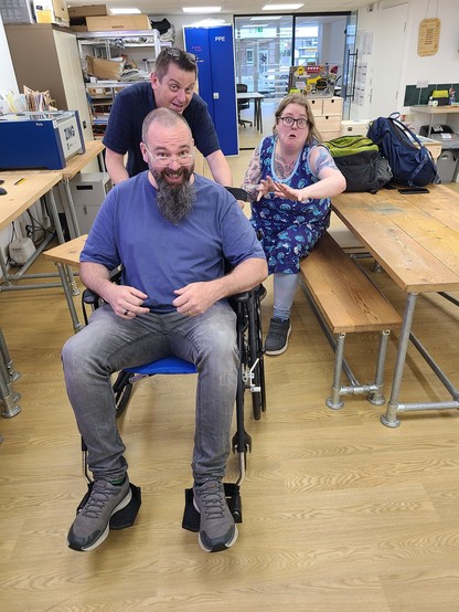 3 white people in a well-lit, open space. The front-most person is shaved bald with a long beard. They're wearing a blue shirt and grey jeans, and sitting in a wheelchair looking happy. The person behind them has short, dark brown hair and is pushing the wheelchair, he is also looking happy. The person furthest behind has light brown hair and is wearing a blue dress. They're sitting on a bench and reaching towards the camera with a despairing expression on their face.