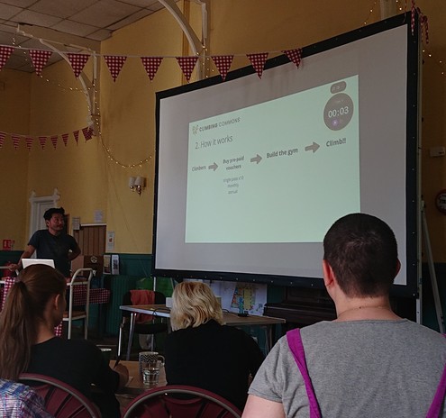 A person presenting a slide which reads: "Climbers ➡️ buy pre-paid vouchers ➡️ build the gym ➡️ climb!!" 