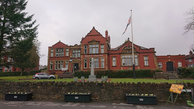 A red brick hall with sandstone detailing sits behind a small lawn which features a war memorial and a flagpole flying the Union Flag