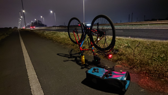It's night time on a lit dual carriageway with a segregated cycle lane running alongside.

A gravel bike is stood upside-down on its saddle and handlebars, and on the ground next to it are some tools, a pannier, and a water bottle.