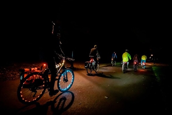 A group of cyclists ride through a park at night.  The rider nearest to the camera has multi-coloured lights wrapped round the frame of their bike and along the rims of the wheels.
