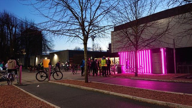 A group of cyclists amass next to a shipping container that's been converted into a bike workshop.  The container is painted black and has stripes of bright pink LED strips running vertically along its length, blazing into the gathering dusk