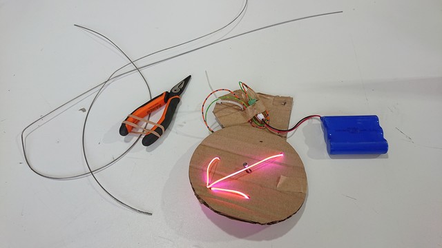 A white desk with a couple of old bike brake cables and some pliers, next to a disc of cardboard with some wires and a battery connected to it.  On the disc there's an arrow made from three lengths (actually it's one length looping round from behind) of illuminated pink LED "noodle"