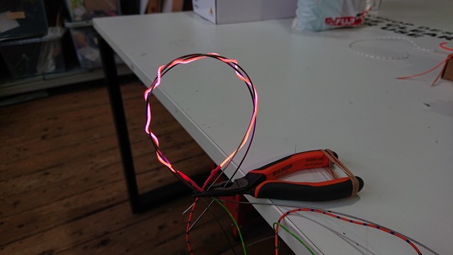 A loop made of plaited LED noodle and bike gear/brake cable, held by a set of long-nosed pliers.  The handles of the pliers are resting on the edge of a table with the nose hanging over the edge.  The loop is vertical in free air, and the LED noodle makes a wavy pattern due to the plaiting.