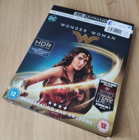 A close-up shot depicts the 4K Ultra HD Blu-ray Digital version of the movie “Wonder Woman” on a wooden surface. The cover features Gal Gadot as Wonder Woman, adorned in her iconic armour and headdress. In the background, a golden, stylized “W” is visible above the movie title, set against a dark blue backdrop with the DC logo in the top left. The text “4K Ultra HD Blu-ray Digital” is printed in silver across the top.

A sticker in the upper right corner displays the price of £2.00. The cover also showcases HDR (High Dynamic Range) technology. A quote from The Sun newspaper, “EXHILARATING AND AWESOME,” is printed near the bottom.

Below Wonder Woman's image, a logo advertises the Justice League movie, which was released in theatres in November 2017. The age rating of “12” is marked on the left and right bottom angles.