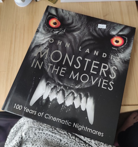 A slightly tilted close-up shot features the book “Monsters in the Movies” by John Landis sitting on a surface and partially on a person's lap or clothing. The book's cover is largely black and white, featuring a menacing monster face with striking orange eyes and large, sharp teeth. The title, “MONSTERS IN THE MOVIES,” is printed in a bold, white font, dominating the central part of the cover, with “John Landis” printed above it. Below the monster's teeth, the subtitle reads, “100 Years of Cinematic Nightmares,” suggesting a comprehensive exploration of horror films.
