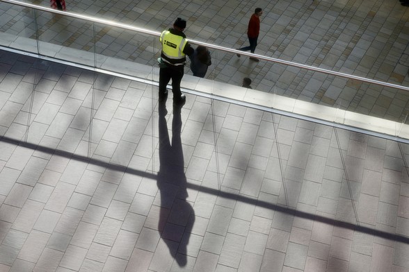 A person leans on a railing, looking down on people walking below. The leaner’s shadow is long and pronounced, bisected by the shadow of the railing. 