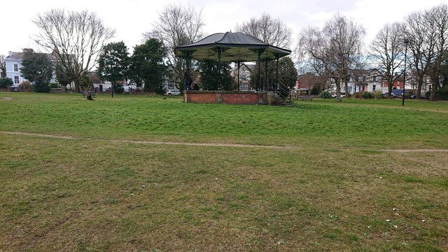 A wrought iron, octagonal bandstand, painted black, on a low red brick base in an open grassy park, with a couple of dirt paths passing by it.

This one is in Newsham Park in Liverpool.
