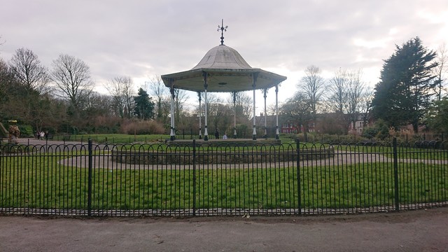 Another octagonal wrought iron bandstand, this one painted white with black details.  It's on a stone-walled pedestal and surrounded by grass and then a black metal fence.

This one in Derby Park in Bootle