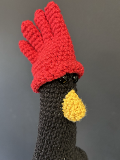 A close up headshot of a crocheted stuffed toy version of Feathers McGraw from Wallace and Gromit, complete with red glove hat and beady eyes
