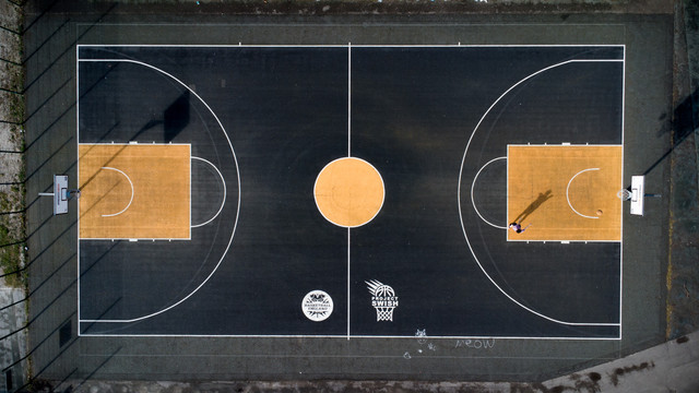 Aerial view of an outdoor basketball court, dark surface with orange rectangular keys, oriented in landscape. On the right hand side, a lone figure shooting the ball can be seen.