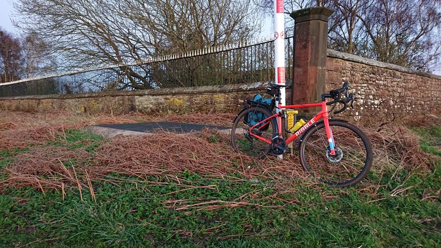 An orange gravel bike with a petrol blue pannier leans against a red-and-white striped pipe (warning of the location of an underground gas pipe), in front of a sandstone wall, topped with iron railings which are painted black with white pointed finials