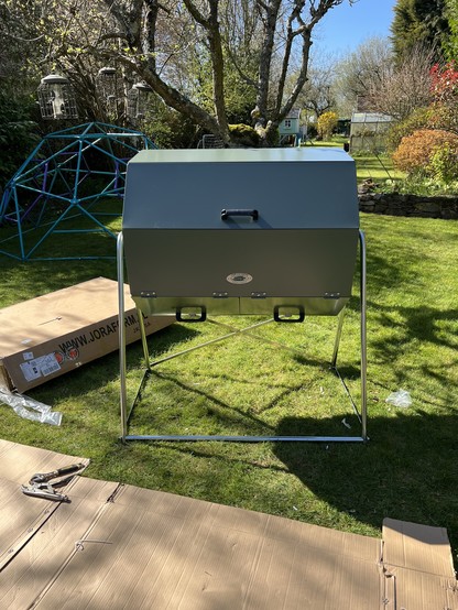 A finished Joraform Big Pig tumbling hot composter amongst its boxes and other packaging, under an apple tree on the grass. 