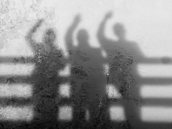 Black and white photo: three people's shadows on water, each person has one arm raised over their head, like they're celebrating. Running from side to side of the image are four parallel straight lines, bars of a wooden fence.
