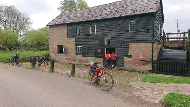 A two storey building built in sandstone and black-painted horizontal planks of wood, with a slate roof.  Attached to the right-hand end of it is a wooden overshot water wheel, also painted black.

A series of wooden bollards mark the edge of the road past the flour mill, and three bikes are leant against them.  Two of the cyclists are stood at the open doorway to the mill, the third was holding the camera.