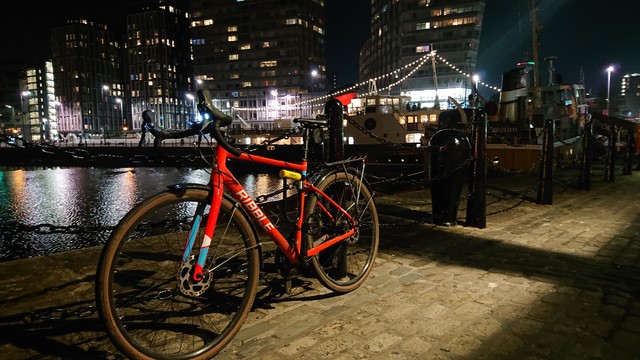An orange gravel bike leans against the railings alongside Canning Dock. The lights of night-time Liverpool reflect off the water, and two ships are moored in the background: tug Brocklebank and former ferry Daffodil, now a restaurant and festooned with strings of white lights 