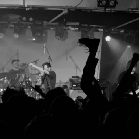 A black and white image of a band on stage, showing the drummer and singer to the left of the image. The bottom of the image shows the silhouetted crowd, and a good portion of the right hand side of the image is taken up with two legs pointing directly upwards. The legs appear to be wearing Dr Marten boots.