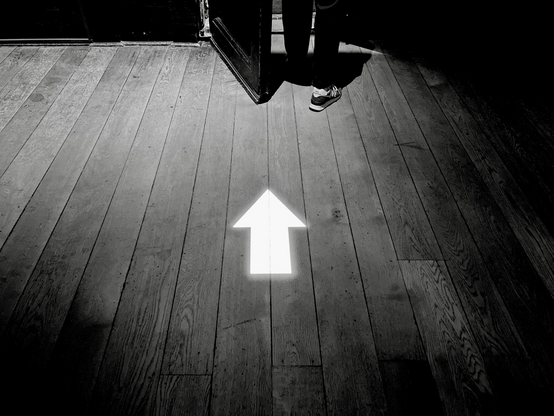 Bw shot of a white arrow projected on a wooden floor in a gloomy room. Where the arrow points, a pair of feet are just disappearing through an open door 