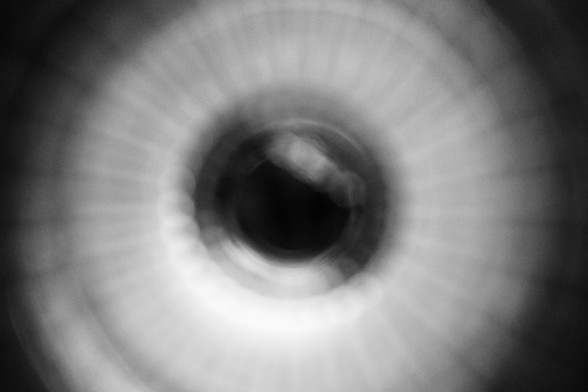 Black and white shot of what looks like a blurry eye: dark pupil surrounded by white inward stripes of pale light. It’s actually a water glass from Ikea sitting on a black granite worktop 