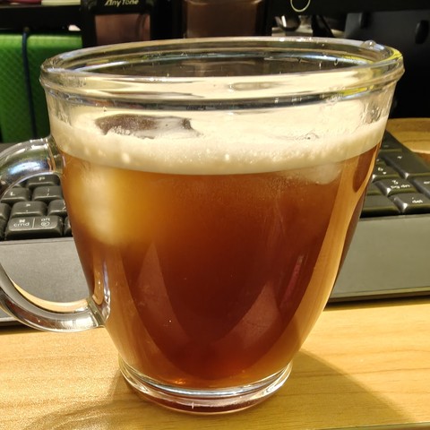 A photograph of an iced coffee in a glass mug.  You can see the ice cubes in the coffee, and a layer of foam on the surface of the coffee itself.  The lighting actually makes the coffee appear lighter/weaker than it actually is, having a light golden appearance.