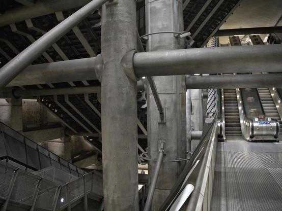 A giant underground space filled with escalators, beams and pillars. The materials are unadorned concrete and steel.