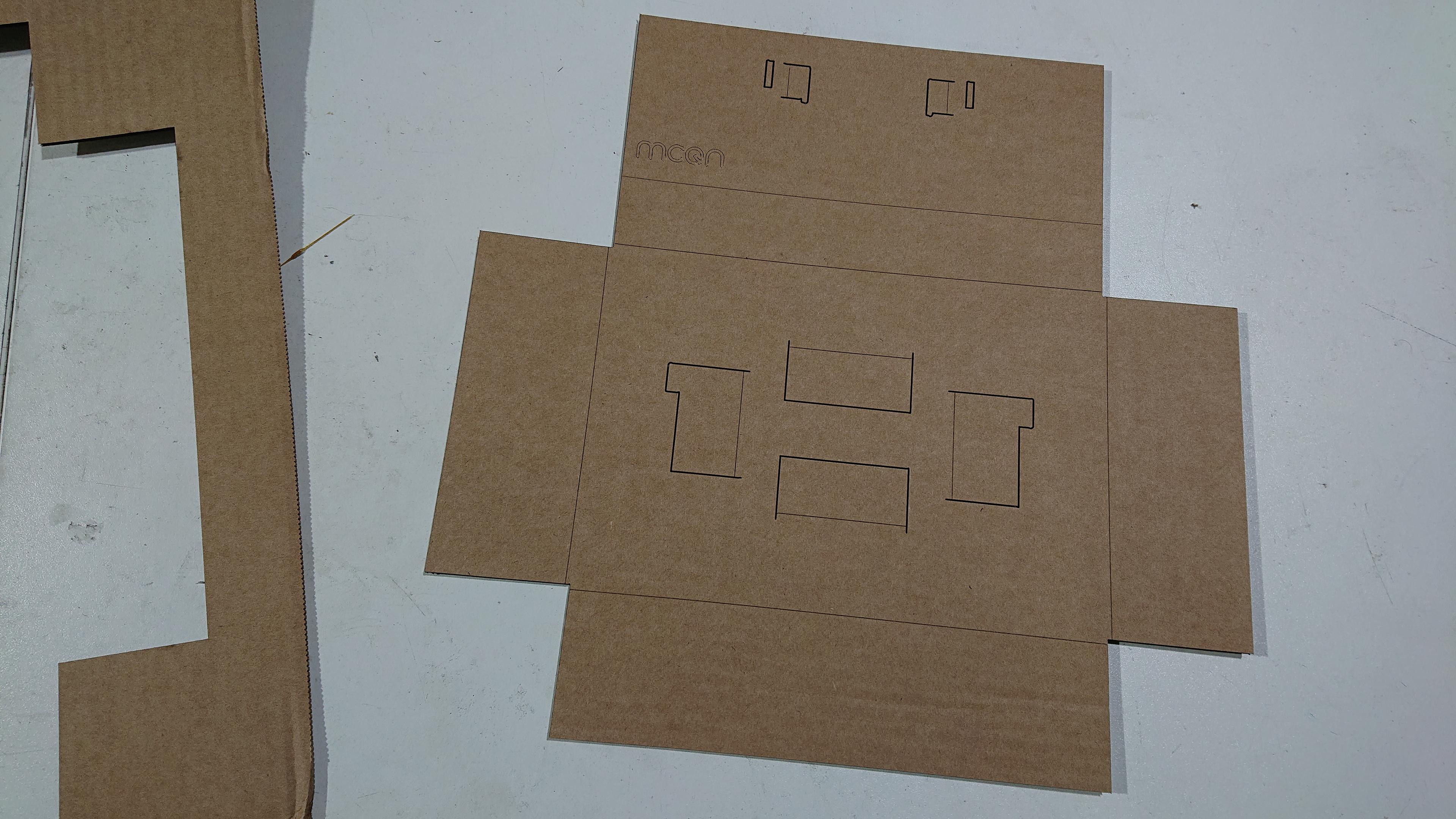An unfolded box insert, laser-cut from brown corrugated cardboard, sits on a white table.  The sheet of cardboard it was cut from can be seen off to the left.

The insert is roughly in a cross-shape, with some three-sided rectangle cuts made into the centre of the cross.  Those will fold up to hold the box the battery goes in.  Three "arms" of the cross will fold down to form a void below the insert where the saddlebag goes; and the final "arm" folds back on itself to sit above the battery box, and has two fold-up tabs that will locate the My Bike's Got LED PCB.