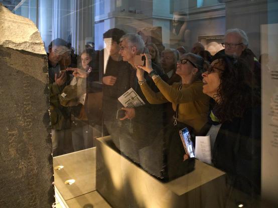 People are looking at the Rosetta Stone in its display case. The picture is taken looking from one side of the Stone, so the people are superimposed on the reflection of the Stone in the front of the case.