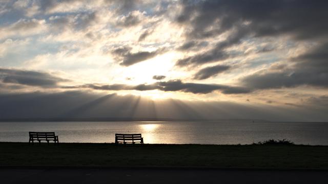 A cloudscape with crepuscular rays. In the foreground two benches are silhouetted.