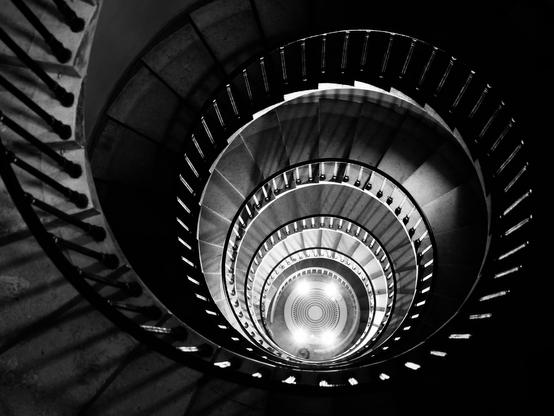 Arty bw photo of a spiral staircase. We’re at the top looking down; there are bright lights at the bottom illuminating everything 